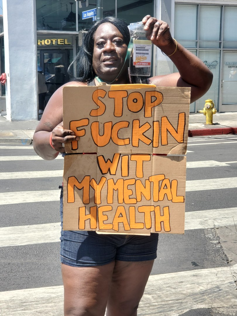 a person holds up a hand-painted cardboard sign that reads "stop fuckin wit my mental health"