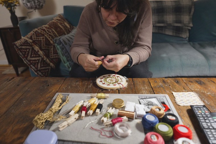 A woman sits on a couch in front of a table and embroiders, embroidery supplies on the table