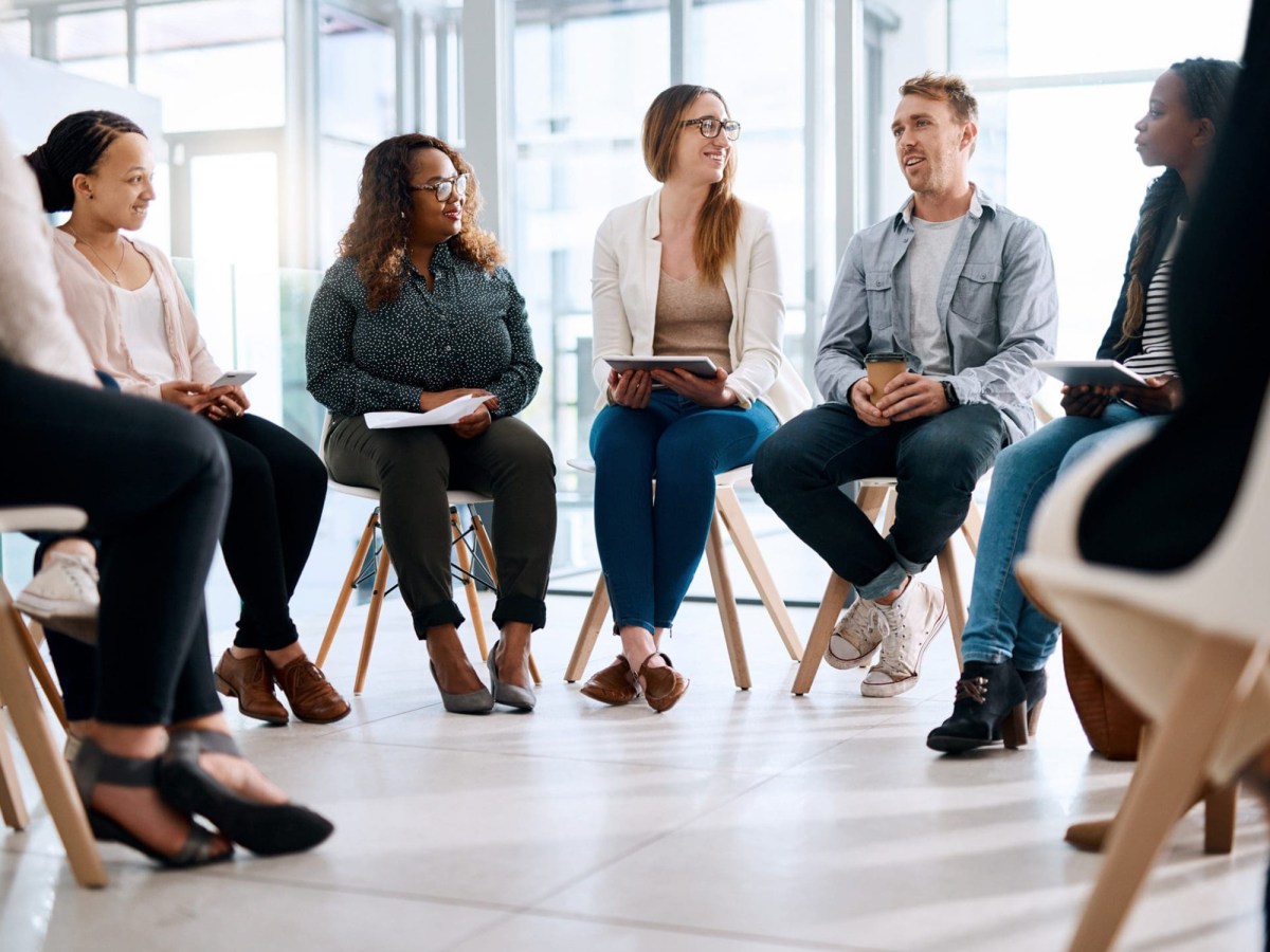 A group of people sit in a circle on chairs indoors, engaged in discussion, with notebooks and coffee cups in hand.