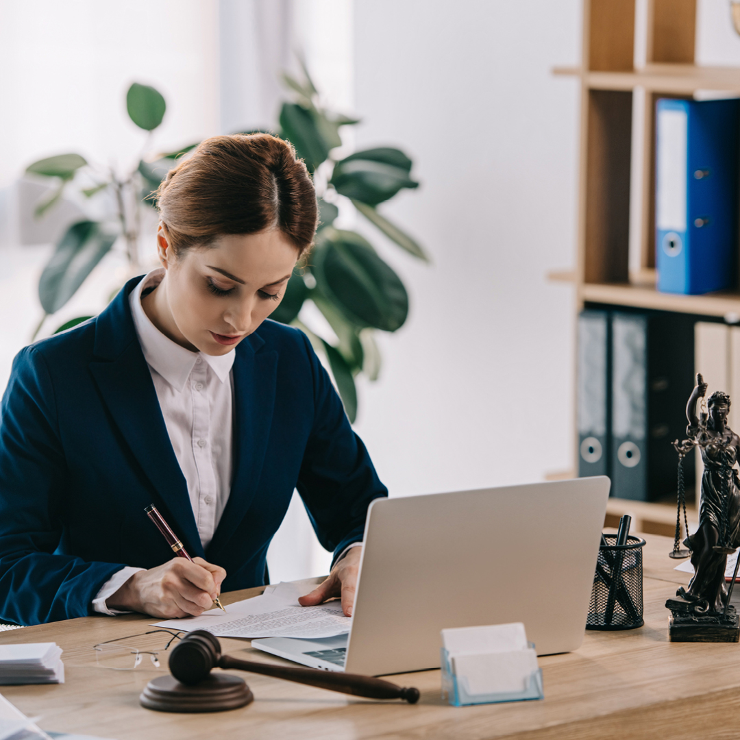 A woman in a suit writing on paper with a laptop, gavel and sounder in front of her