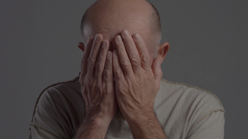 Older man sits with his face covered by his hands, showing distress and exhaustion
