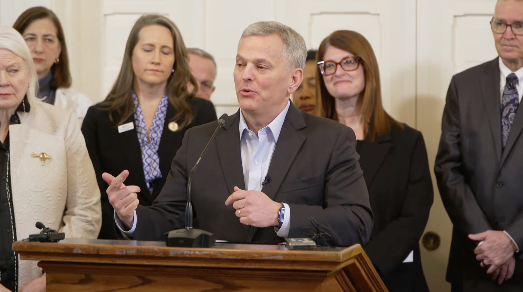 White man with gray hair stands at a podium talking with several people behind him.