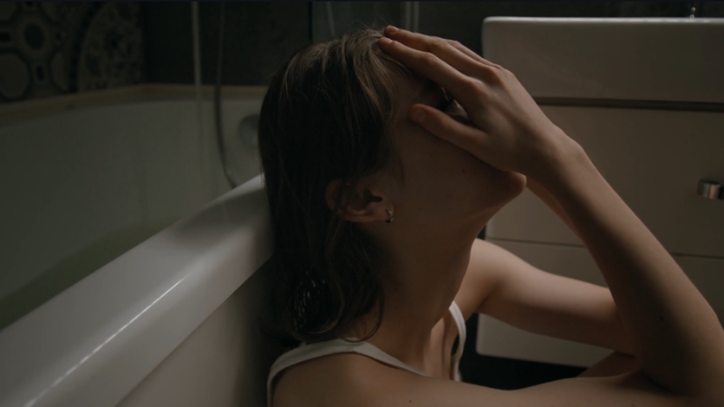 Young woman sits on a bathroom floor with head tilted back and hand over her face, showing emotional distress
