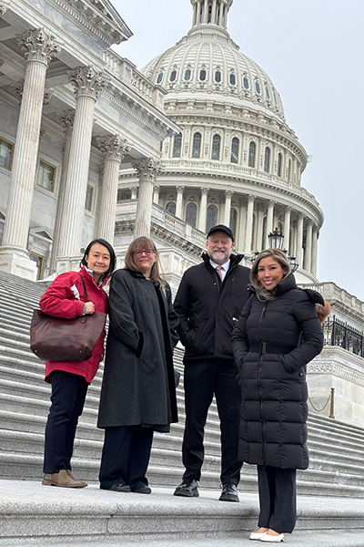 Four people stand on the steps outside the United States Capitol building, dressed warmly, with the dome visible in the background.