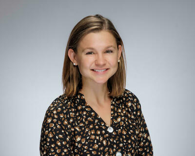 A smiling woman with shoulder-length brown hair and pearl earrings wears a black and brown animal print top.
