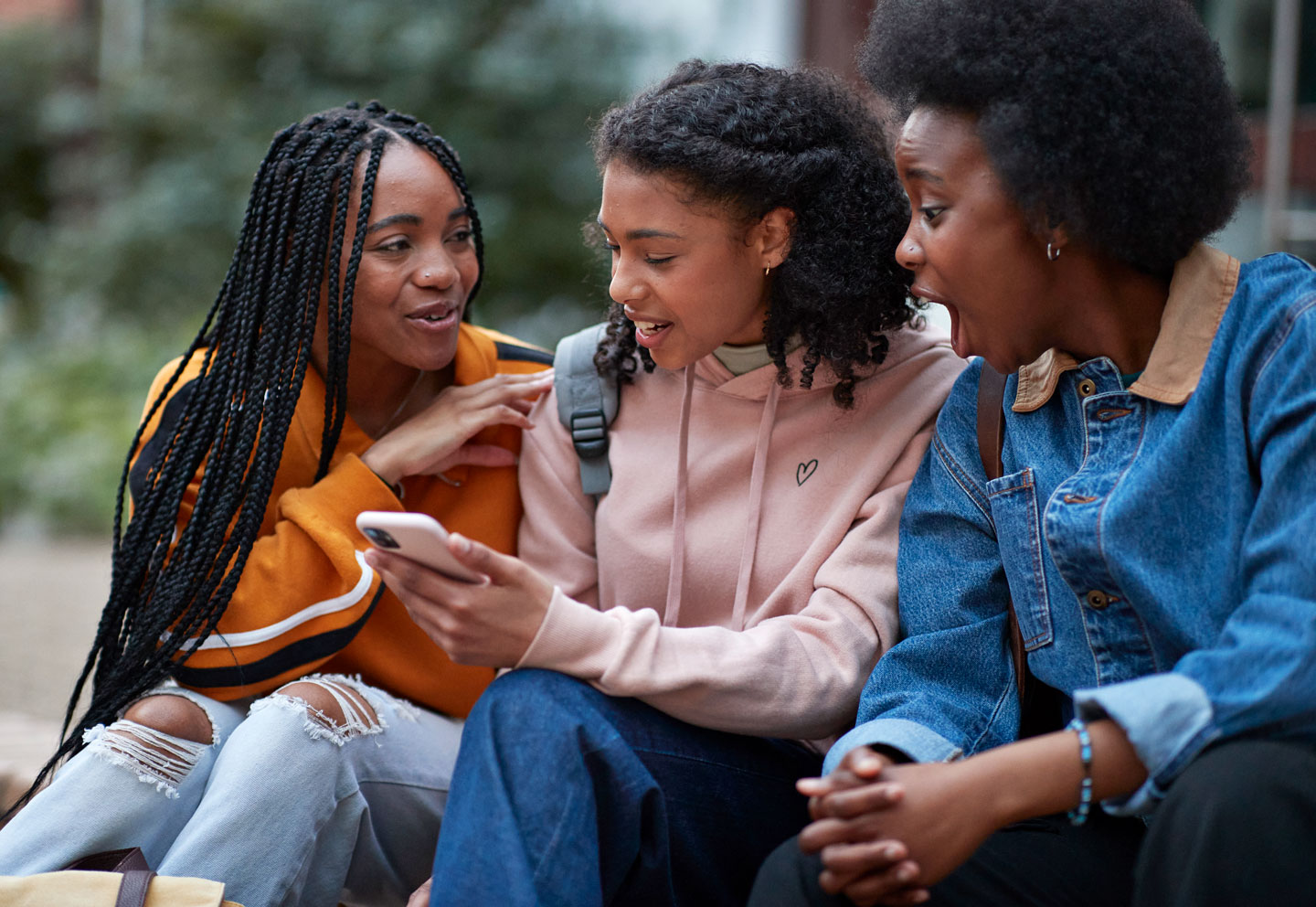three young Black women sit outside together. The middle woman is looking at her phone and talking to her friends. Everyone seems very into the conversation.