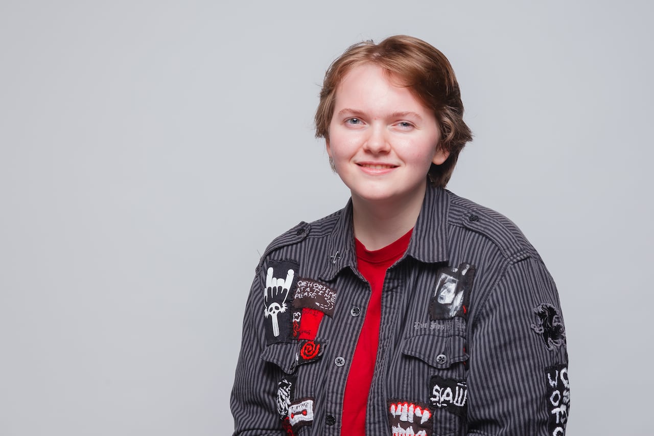 Head and shoulders portrait of young woman with short, brown hair. She is wearing a black and grey striped button down shirt with patches on it. 
