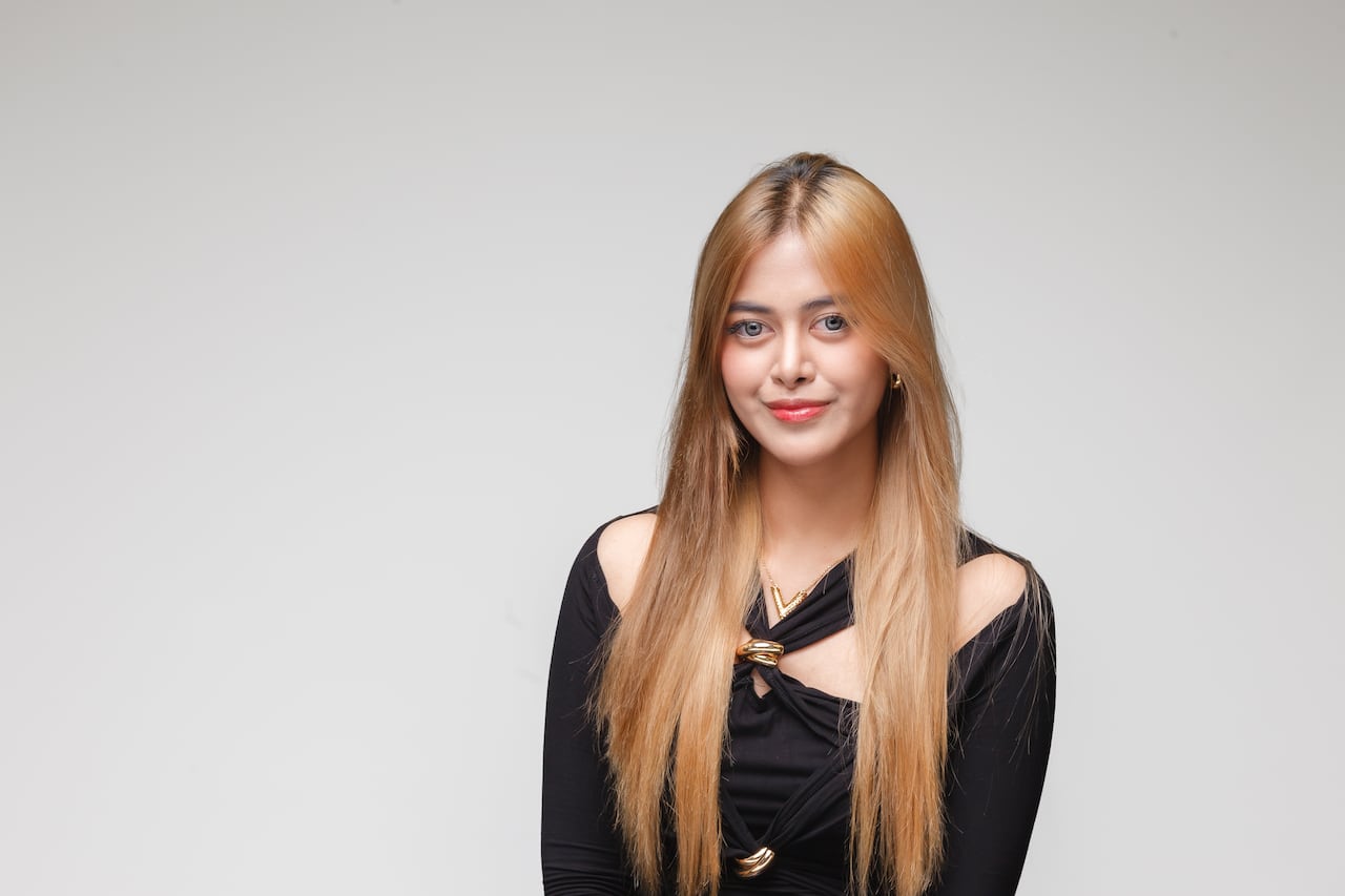 Head and shoulders portrait of young Filipino woman with long, blond straight hair. She is wearing a black top with gold ring accents. 