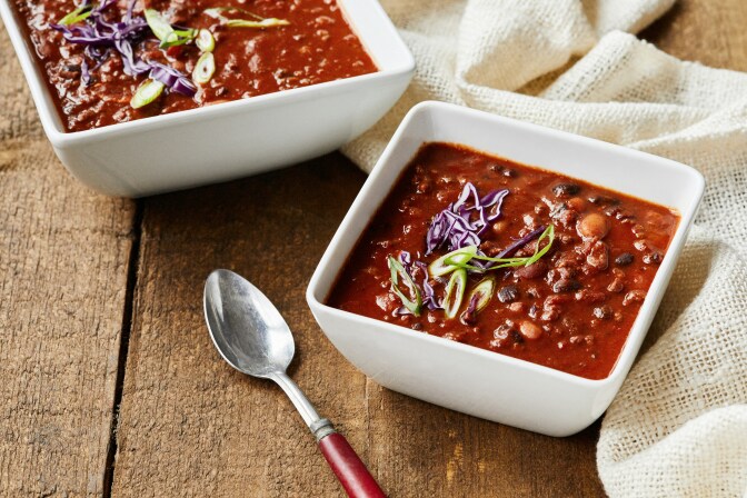 Two square bowls of brownish-red chili with a silver spoon next to one. 