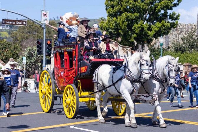 Two white horses pull a red carriage with yellow wheels with several people in cowboy hats riding on top. 