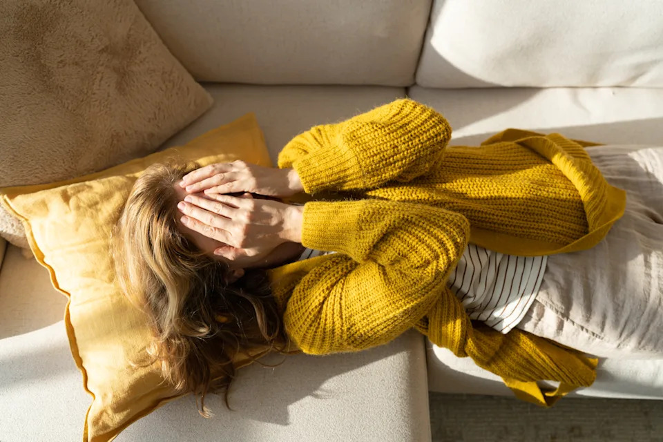 A person lying on a couch, wearing a cozy sweater, hands covering their face