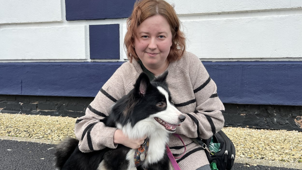 A woman in a striped black and beige jumper crouches with her black-and-white dog on a path outside a building.