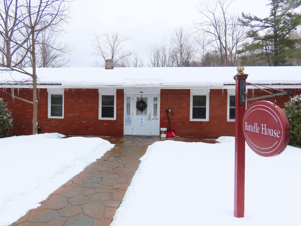 A brick building with snow on the roof and ground, a wreath on the door, and a red sign reading "Battelle House" at the entrance.