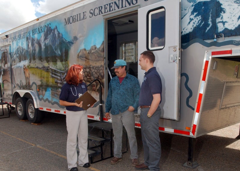 Two staff stand with a migrant worker in front of a mobile screening trailer.