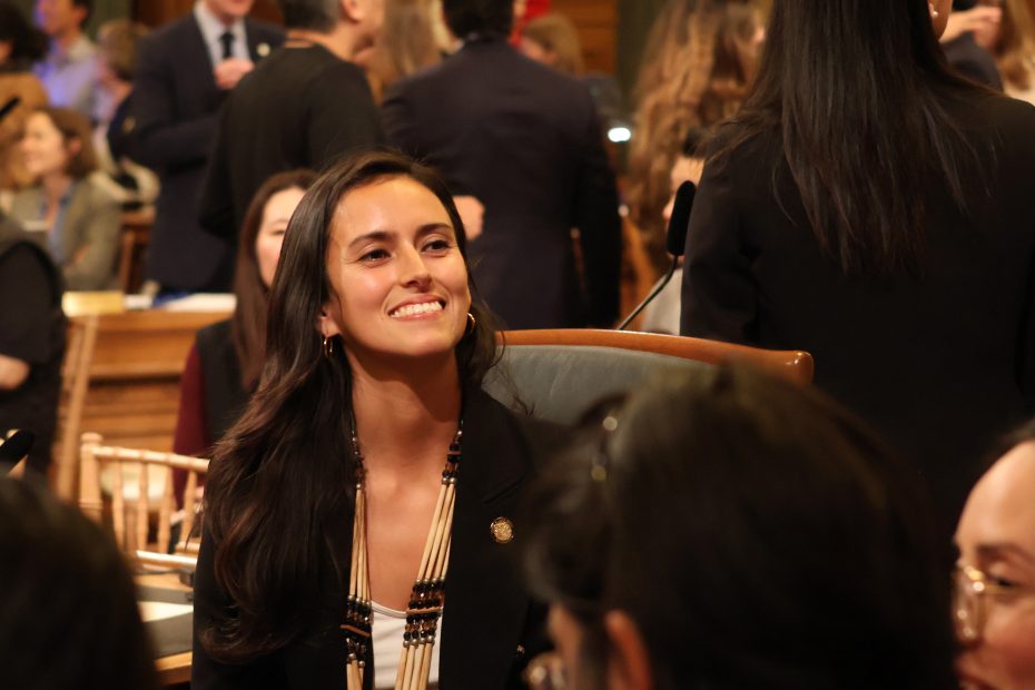 A woman seated at a conference or meeting, surrounded by other attendees, smiles as she looks towards the front.
