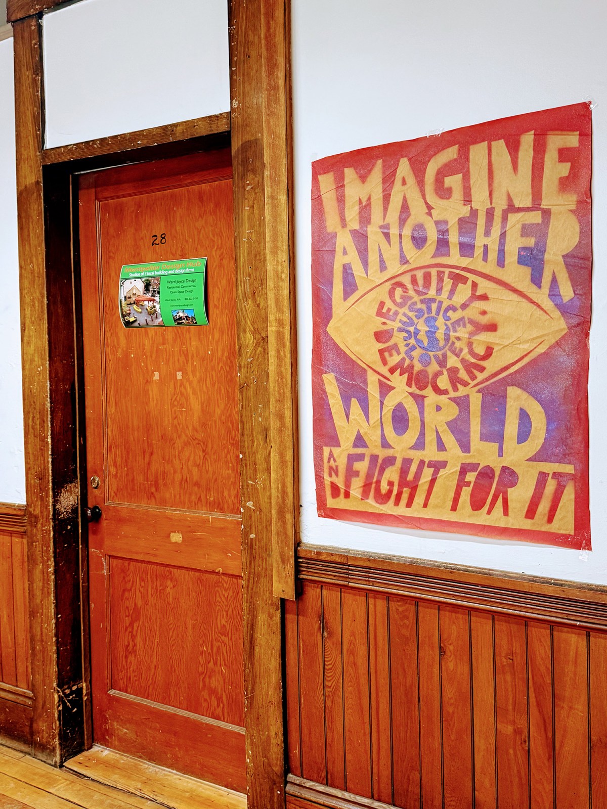 A closed wooden door labeled "2.8" next to a large sign reading "IMAGINE ANOTHER WORLD AND FIGHT FOR IT" with the words equity, justice, and democracy inside an eye graphic.
