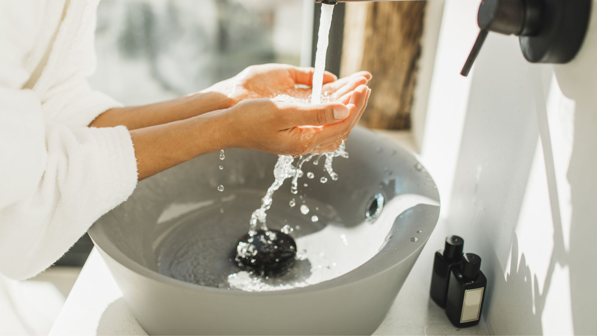 A woman washes her face by hand over a stylish sink in a brightly-lit bathroom.