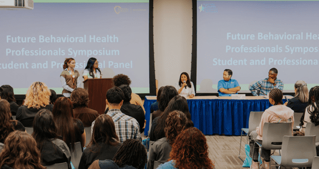 A panel of five speakers addresses an audience at the Future Behavioral Health Professionals Symposium, with event details projected on screens behind them.