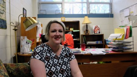 frienly looking woman looks at the camera, sits in front of a desk with papers on it.