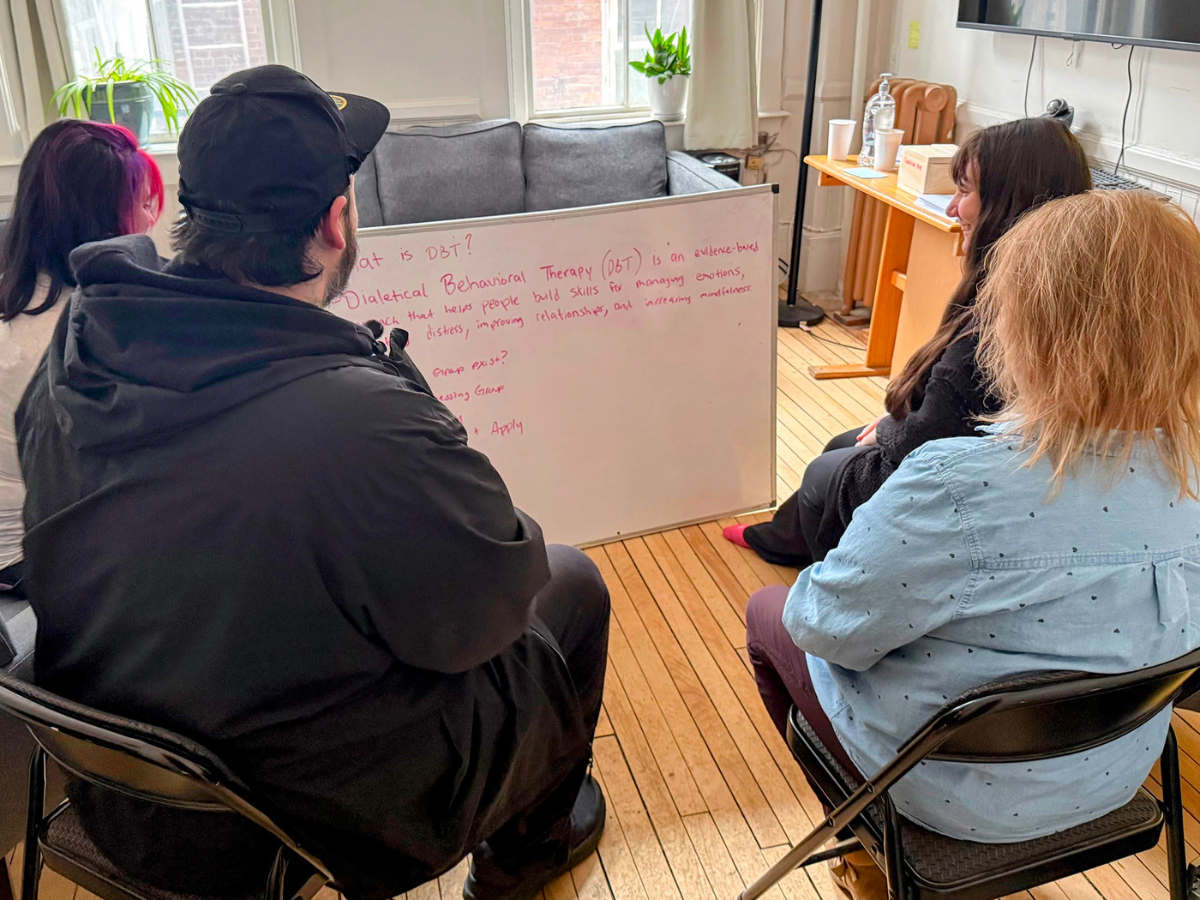 Four people sit in a circle facing a whiteboard with notes about Dialectical Behavioral Therapy in a well-lit room.