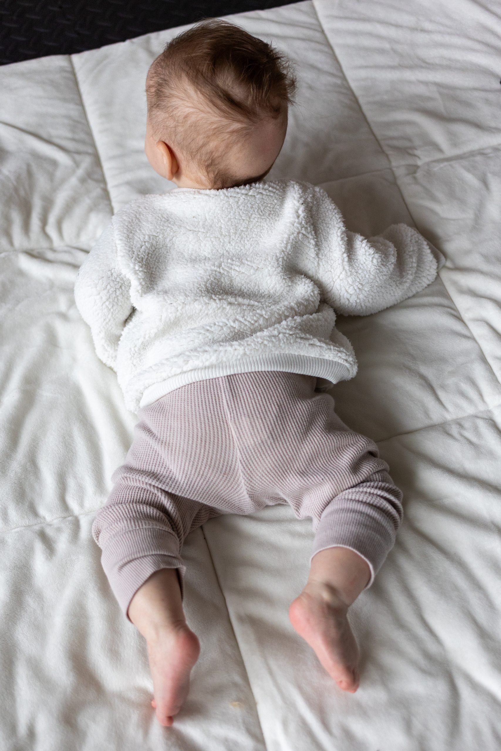 A baby lies on their stomach on a white quilted blanket, wearing a white sweater and light-colored pants.