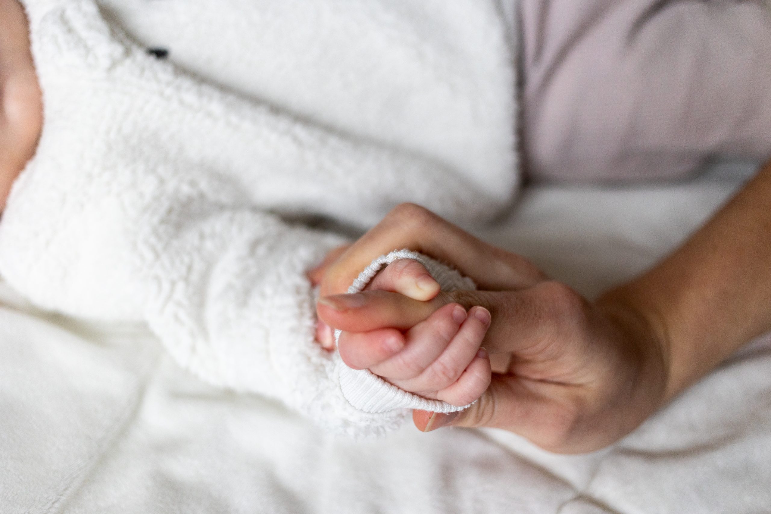 Close-up of an adult hand gently holding a baby's hand, both resting on a white surface. The baby is wearing a white, textured outfit.