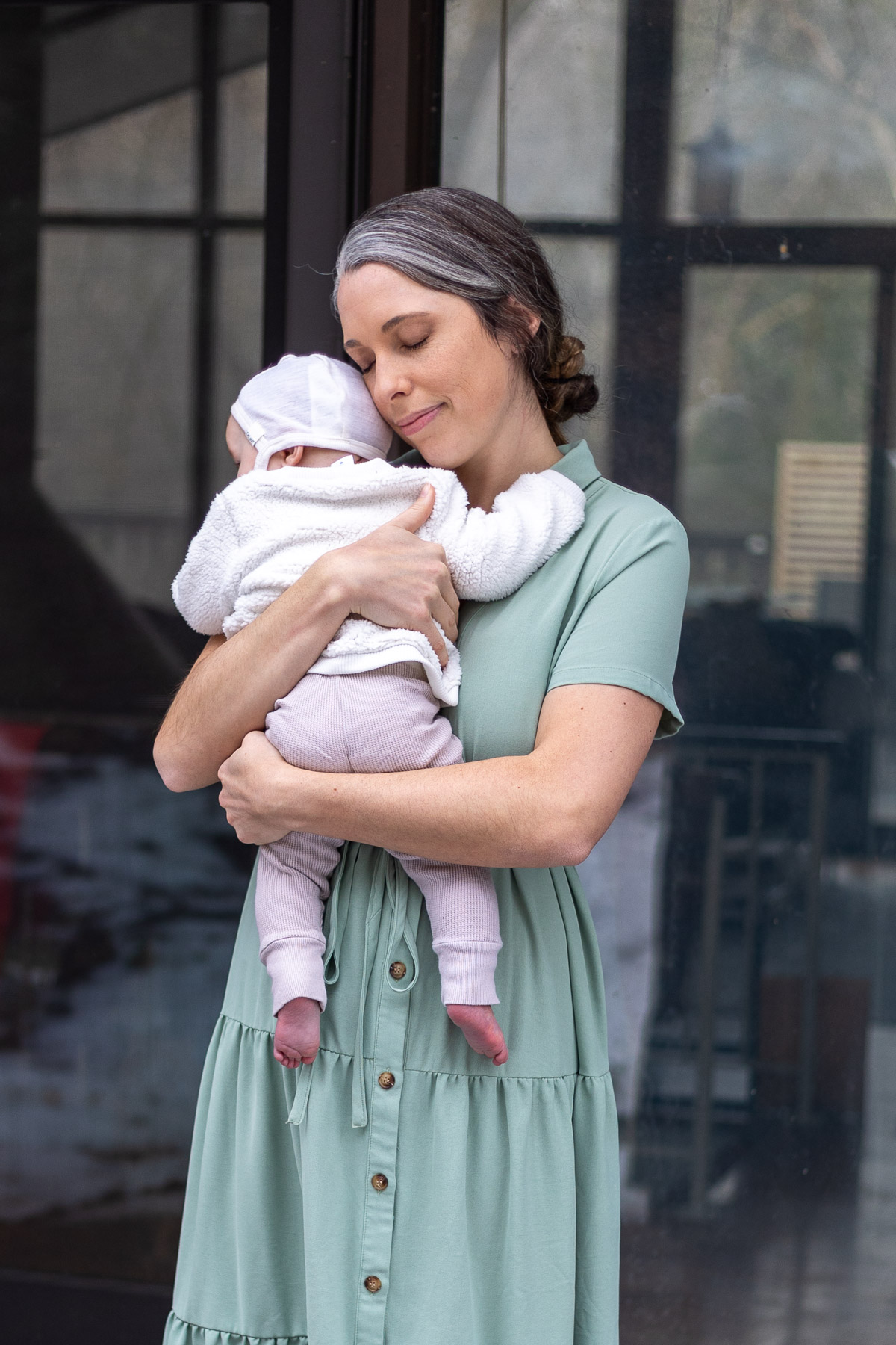 A woman in a light green dress holds a baby dressed in white and pink, standing in front of a glass door.