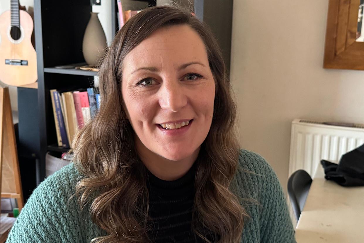 A woman wearing a teal green cardigan with long brown hair looks directly into the camera and smiles. There is a black book case and guitar in the background.