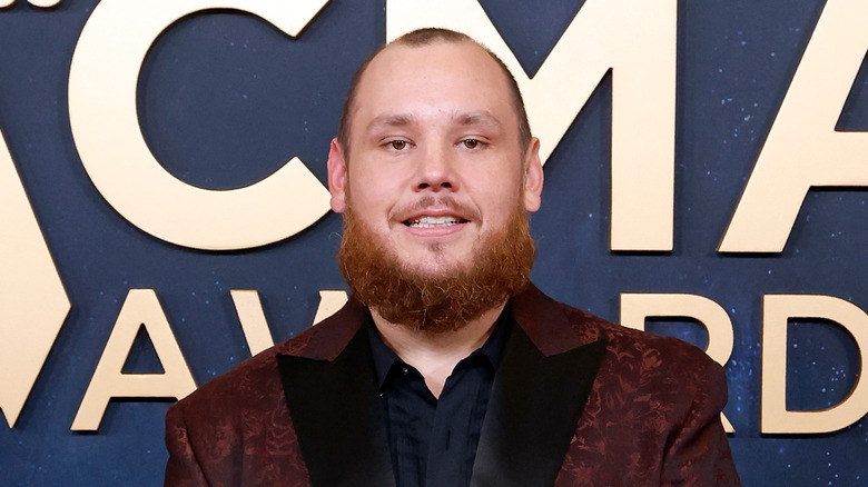 Luke Combs in a red suit coat in front of the ACMA Awards logo