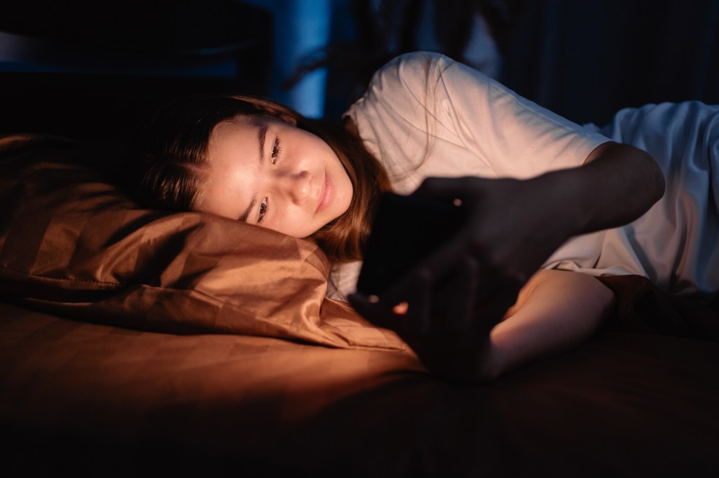 A teenage girl lying in bed, using a phone in the dark, with the phone's screen illuminating her face.