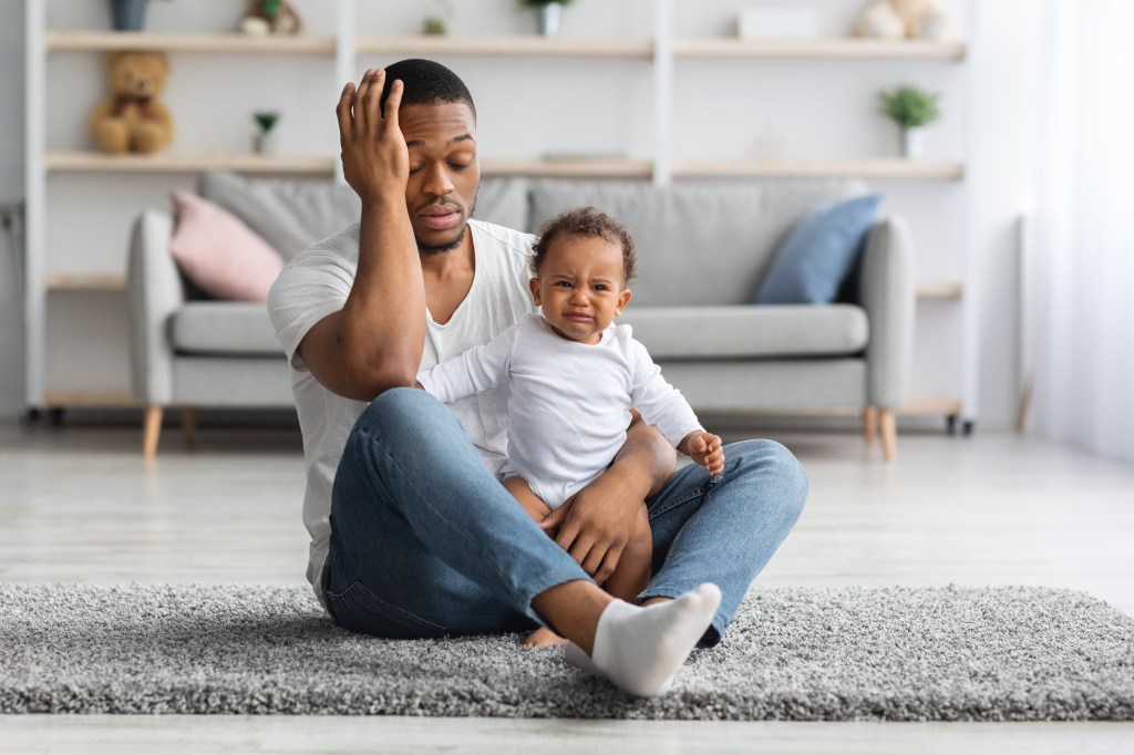 Exhausted Black father sitting on the floor with a crying baby.