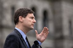 A man in a dark suit speaking outdoors with his hand raised, seen in profile