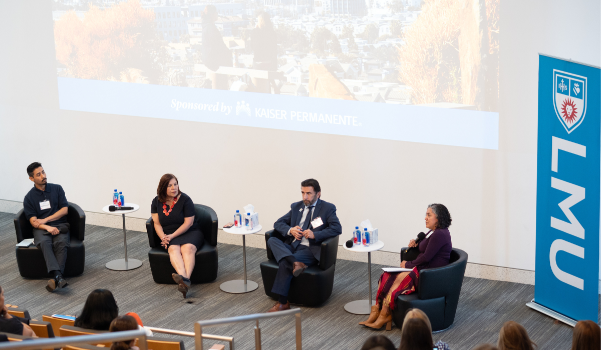 two women and two men sitting in leather chairs in front of an audience with white tables between them and an LMU banner to the right of them.