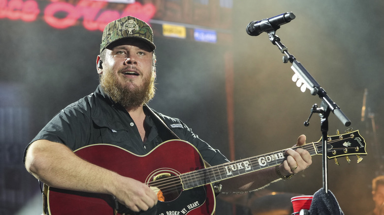 Luke Combs with a guitar behind a microphone on stage.