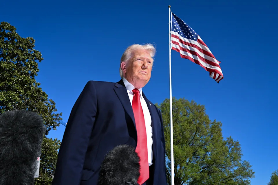 A man in a suit with a red tie stands solemnly in front of an American flag and trees, with microphones visible in the foreground