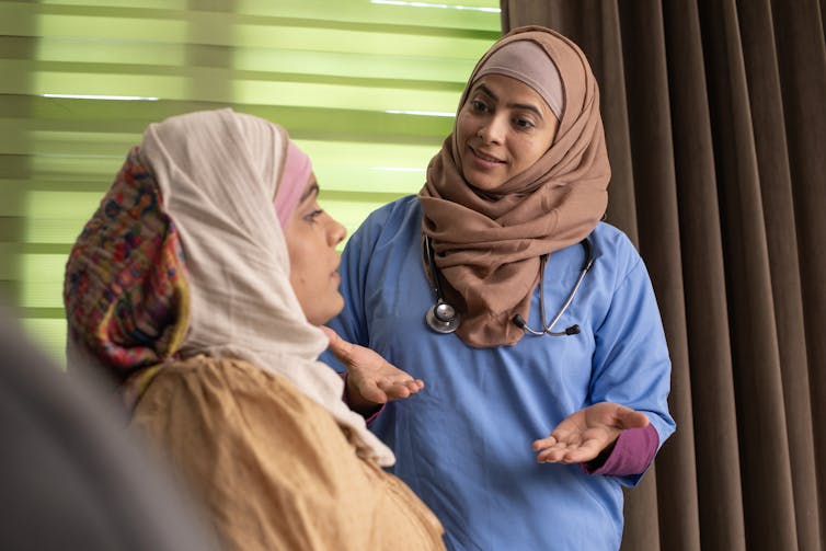 A woman wearing a headscarf speaks with another woman reclining on a bed, who is also wearing a headscarf.
