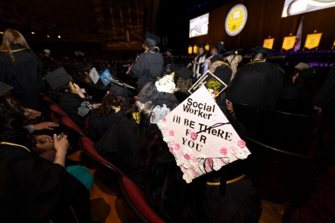 A group of graduates are picture from behind, sitting in an auditorium. A person wears a mortarboard decorated with white and pink flowers and the words, "Social Worker I'll be there for you."