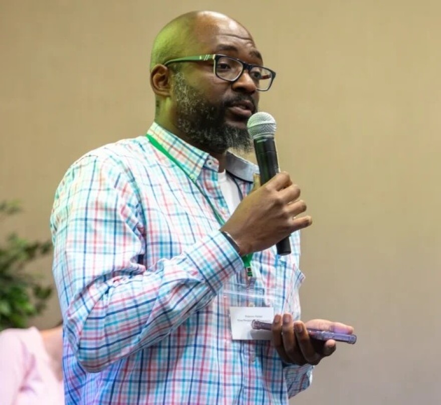 Black bald man with glasses in dress shirt holding microphone and cell phone speaking into the micropone 