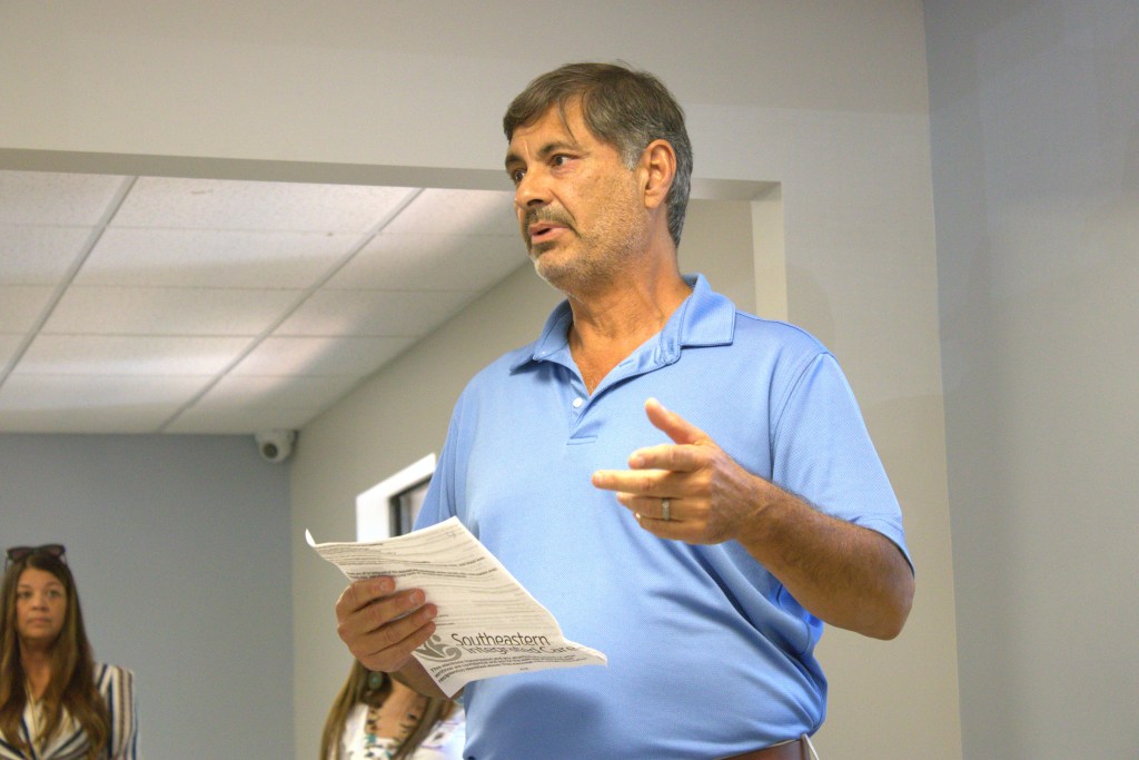 A man in a blue polo shirt speaks indoors while holding a sheet of paper, gesturing with his hand as he addresses a small group. Two people stand blurred in the background, listening.
