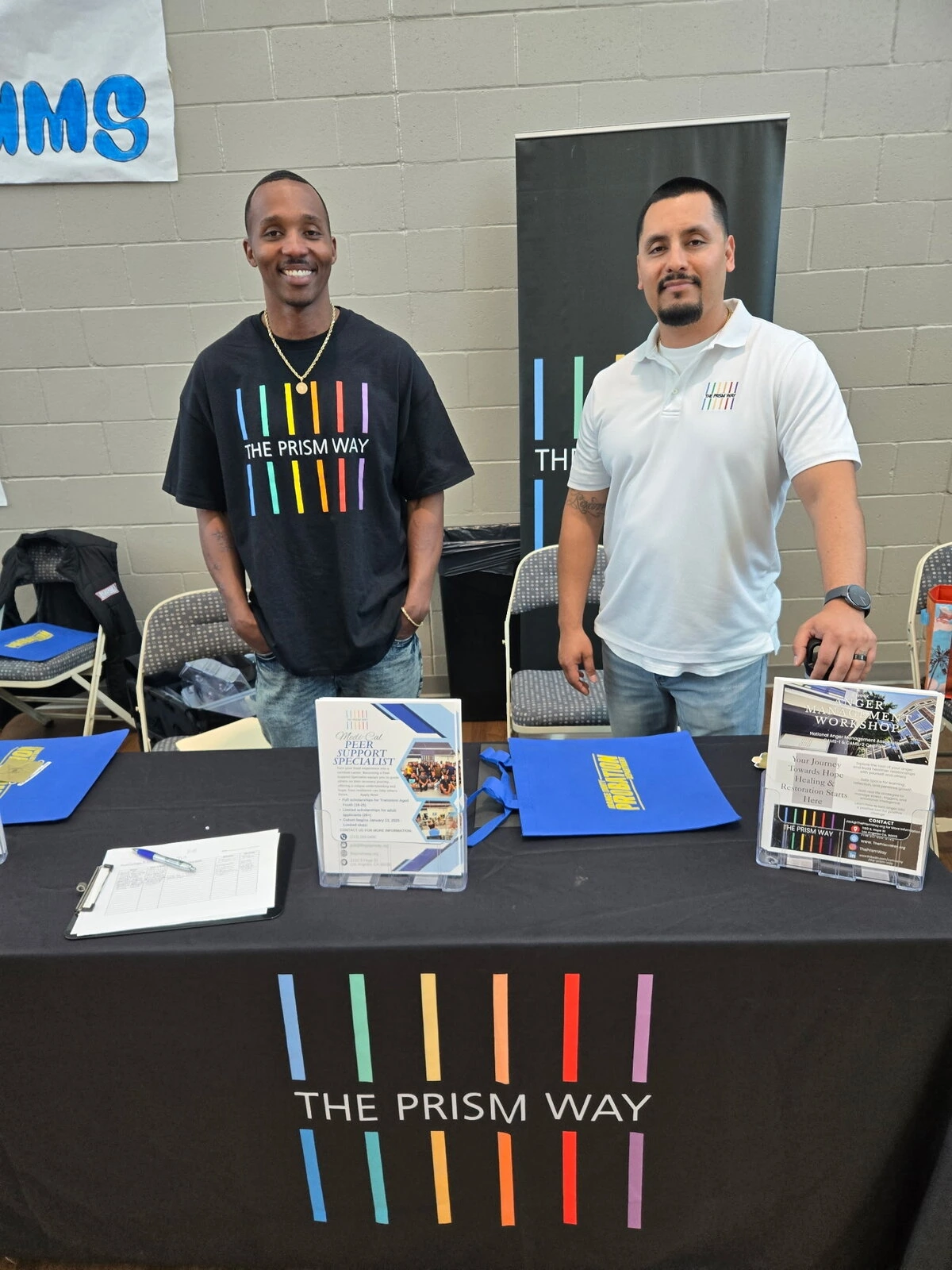 Two men wearing Prism Way shirts stand behind a table with a Prism Way table cloth and other materials.