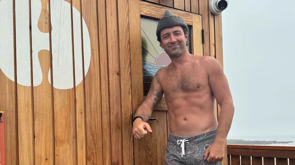 This image shows William Jenkins stood outside a wooden style sauna, he's shirtless but wearing grey pattern shorts and a sauna hat. He has stubble and some tattoos on his arm.