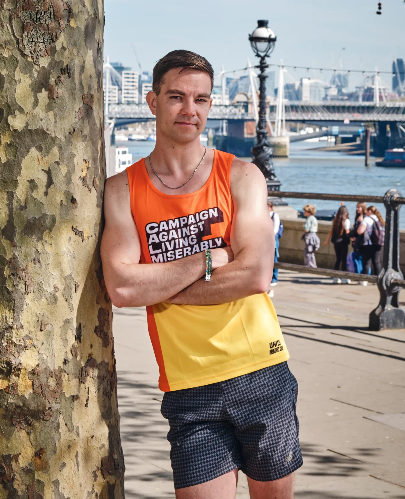 Aubrey Allegretti standing with arms crossed, leaning against a tree, wearing an orange and yellow "Campaign Against Living Miserably" vest.