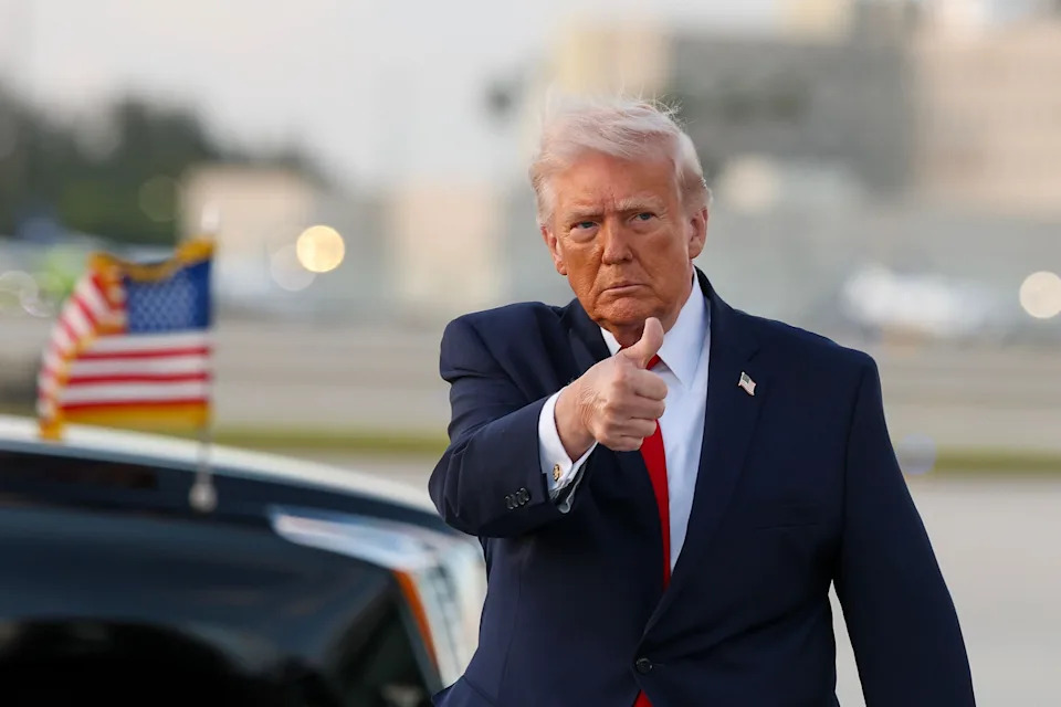 A man in a suit gestures a thumbs-up beside a vehicle with the U.S. flag. Background shows an urban setting and blurred lights