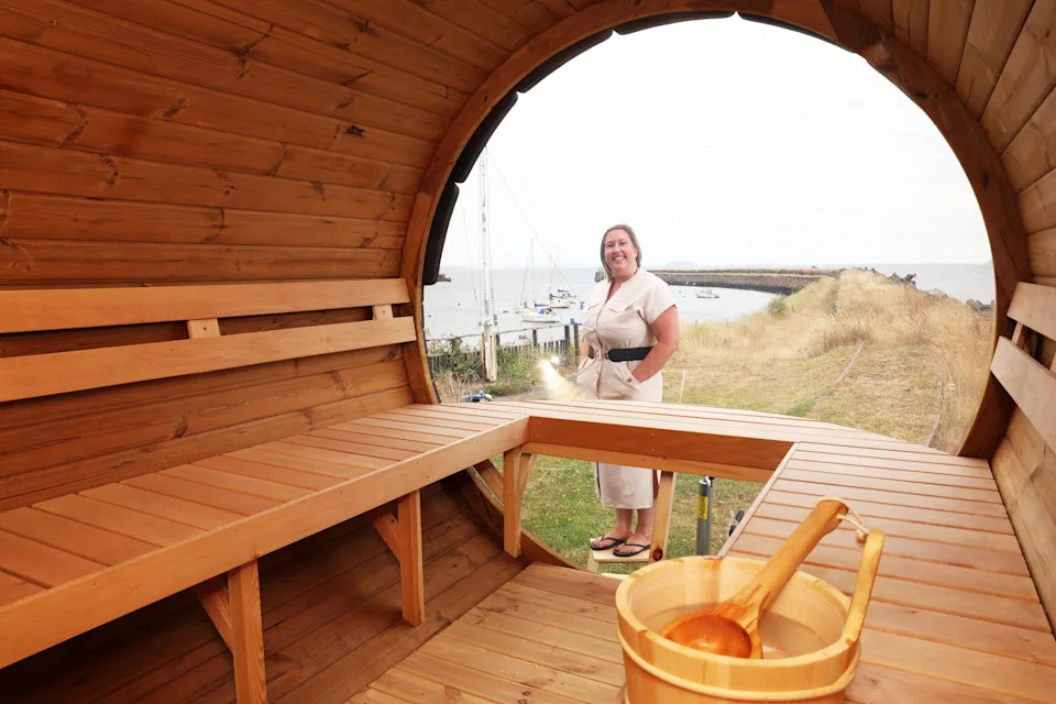 The image shows the inside of a wooden style sauna, Kathryn Donavan, the owner of the sauna is stood just outside. She's wearing a beige dress with a black belt and a pair of black flip flops. She has short blonde hair.