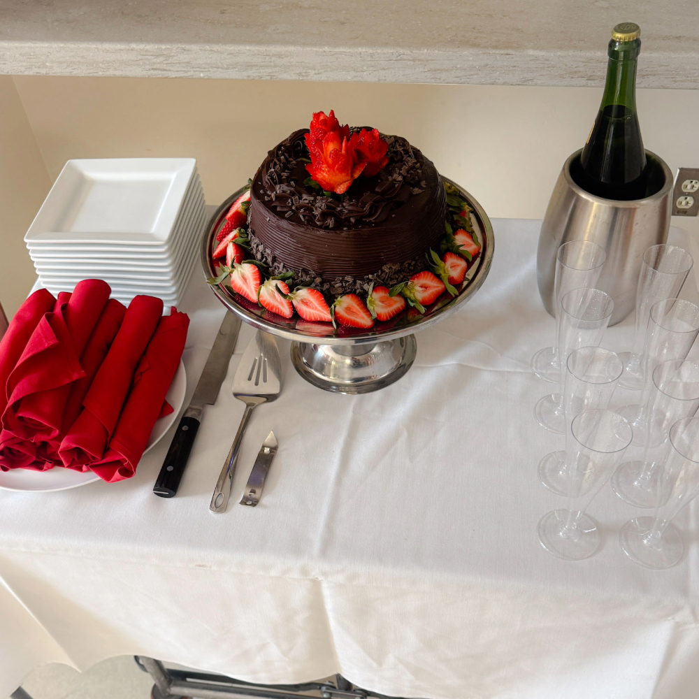 A chocolate cake with strawberries sits on a table alongside champagne, glasses, plates and red napkins