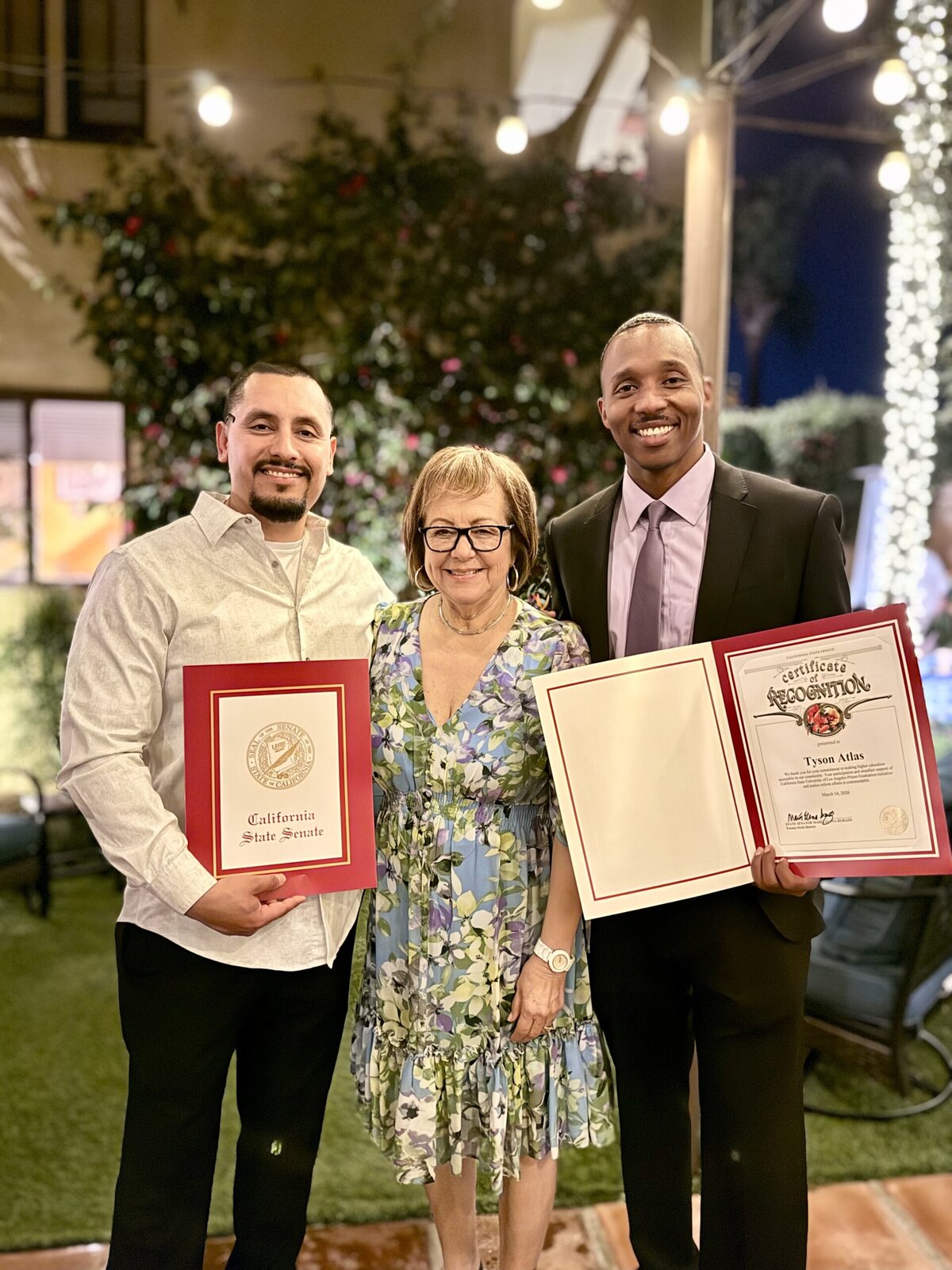 Two men pose with a woman. The men hold up certificates of recognition from the California senate.