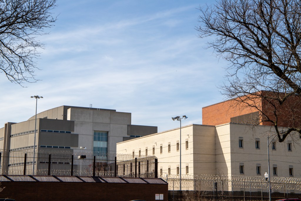 A photo of Cook County Jail from an angle. It is a white building with a barbed wire topped fence in the lower frame.