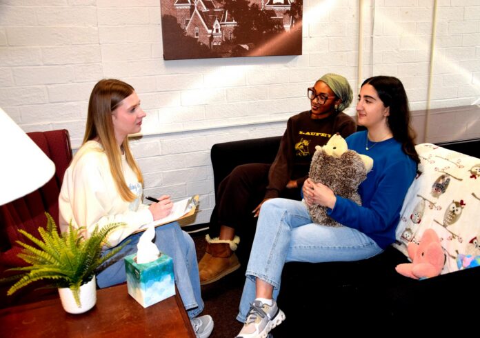 Three women sit in a cozy room talking; one takes notes, while another holds a stuffed animal on the couch.