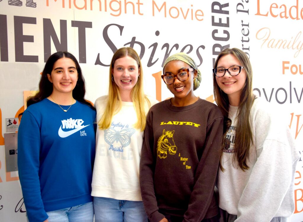 Four young women stand side by side, smiling in front of a wall with inspirational words and phrases.