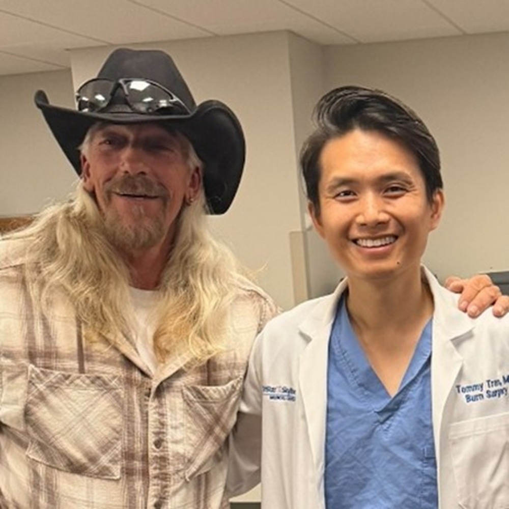 Dr. Tommy Tran, wearing scrubs and a white coat, stands next to patient Bruce Hill, who is wearing a cowboy hat and sunglasses, both smiling in a hospital room